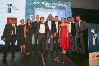 A group of men and women in black tie wear stand on a stage , a brown skin-toned man in in the forfront holding the award highin front of a screen giving the award name - Development of the Year.