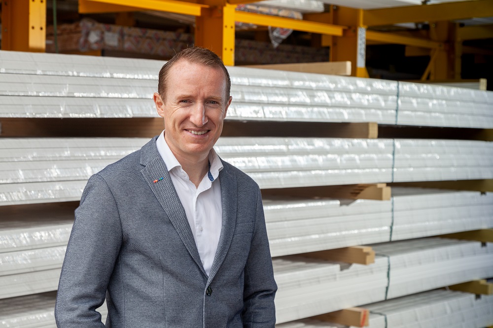 a white man of approximate middle age faces the camera in a casual suit with timber pallets behind him.