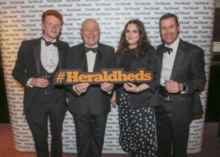 from left a young man with red hair in black tie, an older bald man in black tie, a young women in formal dress and a middle-aged man in black tie hold a sign saying hashtag heraldheds in front of a white backdrop.