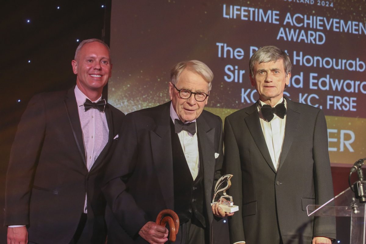 three white men in black tie stand on a stage with the elderly man in centre accepting an award.