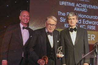 three white men in black tie stand on a stage with the elderly man in centre accepting an award.