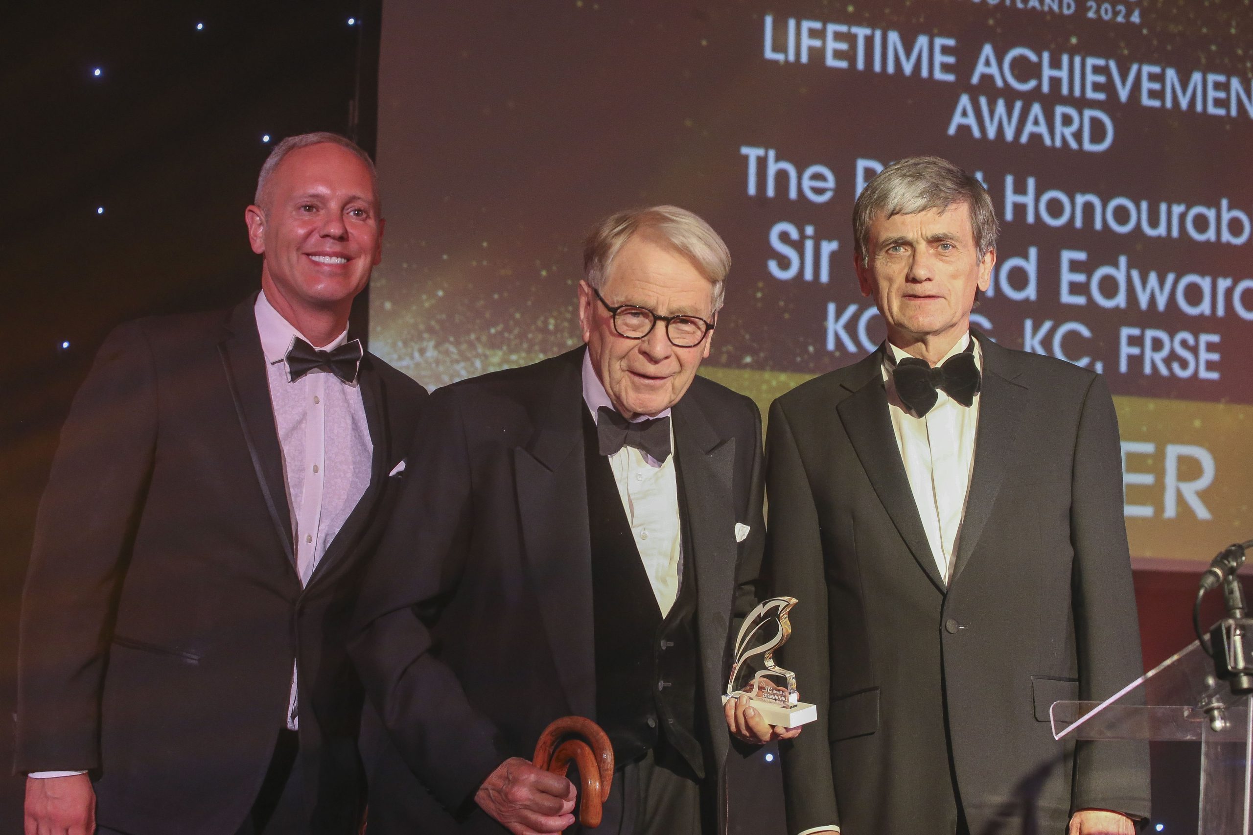 three white men in black tie stand on a stage with the elderly man in centre accepting an award.